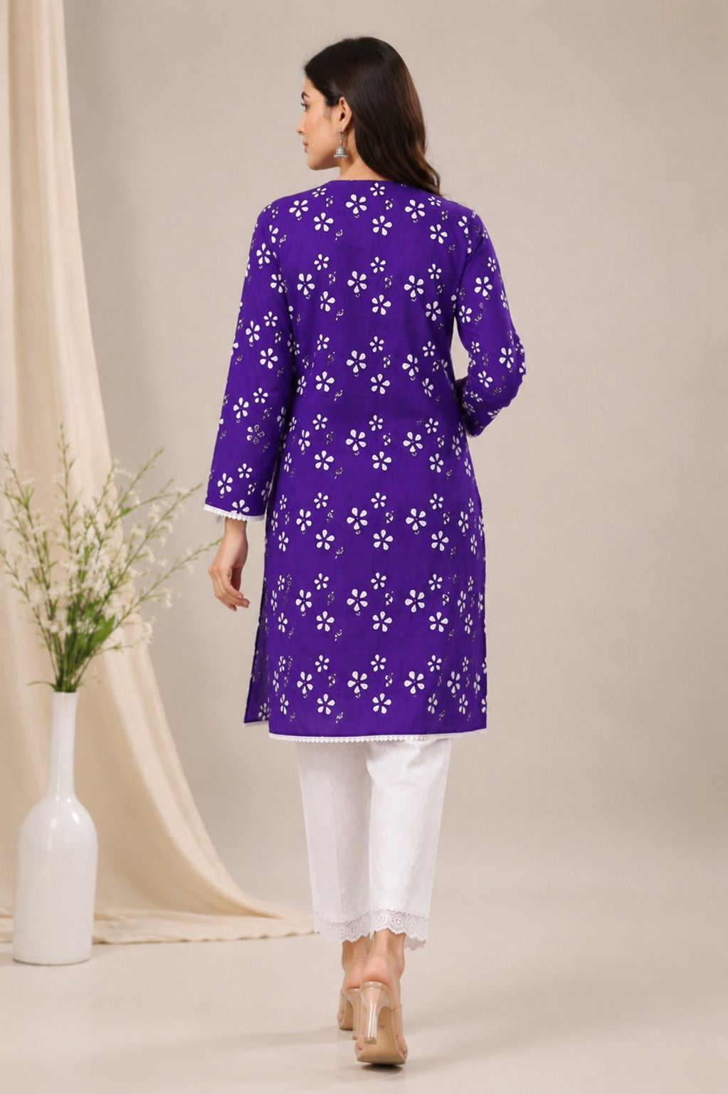 A woman shown from the back wearing a purple long kurta with a white floral print, paired with white pants and beige heels, in a studio setting with a beige backdrop and a vase of flowers.